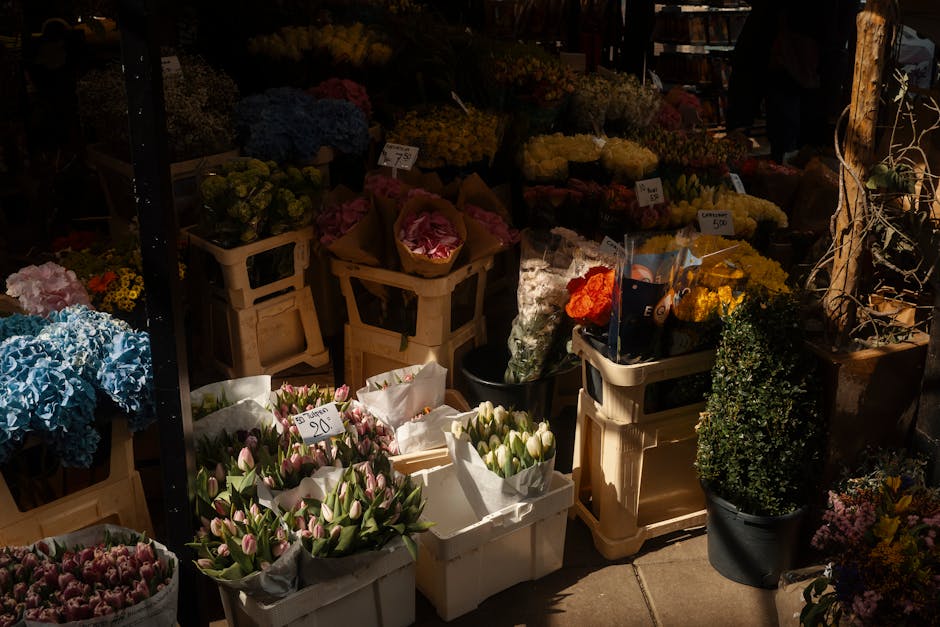 A vibrant outdoor flower market display featuring an assortment of fresh, colorful flowers arranged in white paper wraps and in black and beige crates, including pink and white tulips, blue hydrangeas