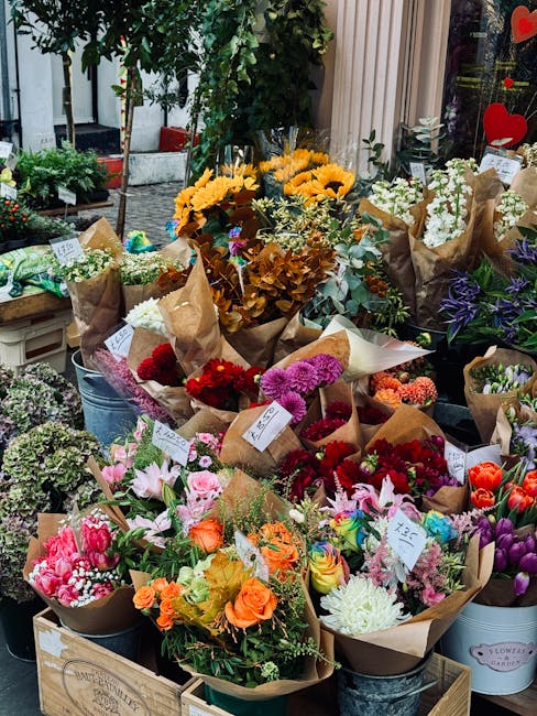 A vibrant outdoor display of various floral arrangements in bouquets and buckets, featuring a wide array of fresh flowers such as bright yellow sunflowers, pink lilies, red and purple carnations, oran
