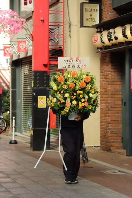 A person dressed in black holding a large floral bouquet arrangement on the street in front of a building with brick and black exterior walls. The bouquet features vibrant yellow, orange, and green fl
