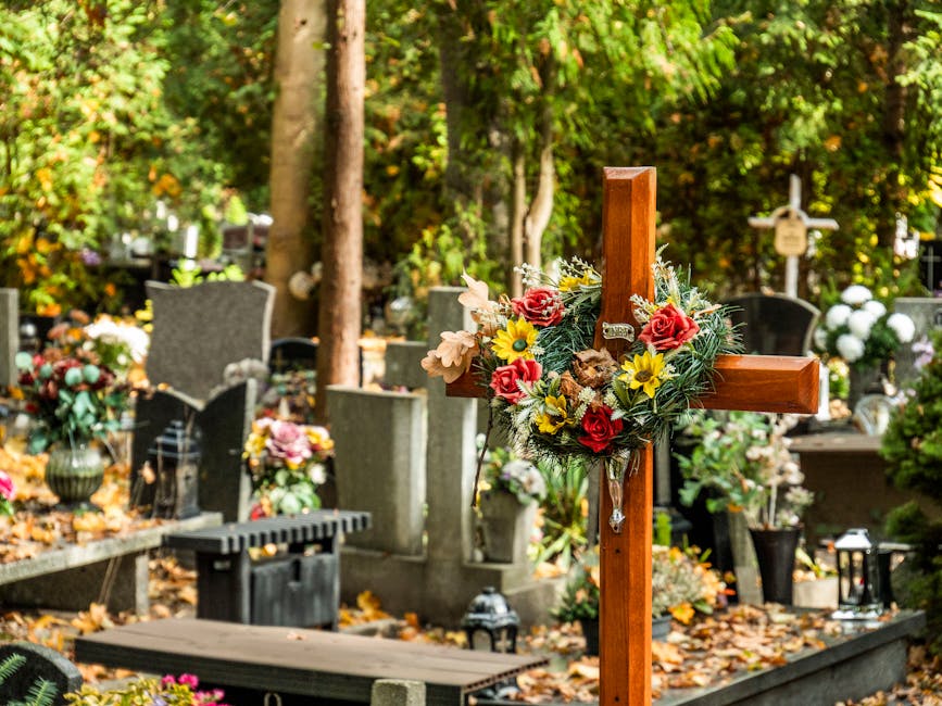 A wooden cross adorned with a floral arrangement features red roses, yellow daffodils, white lilies, and green foliage, situated among gravestones in a cemetery surrounded by tall trees with fallen le